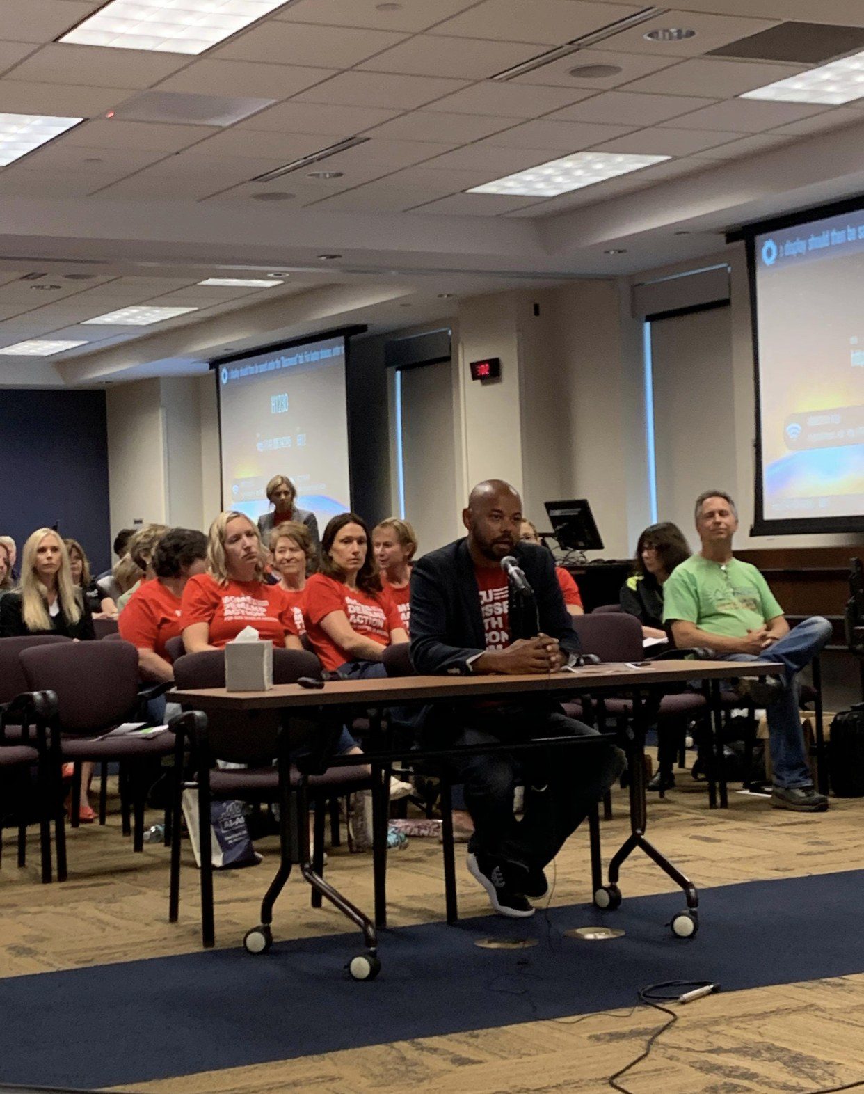 Khary testifying at a hearing table while supporters in red shirts sit behind him